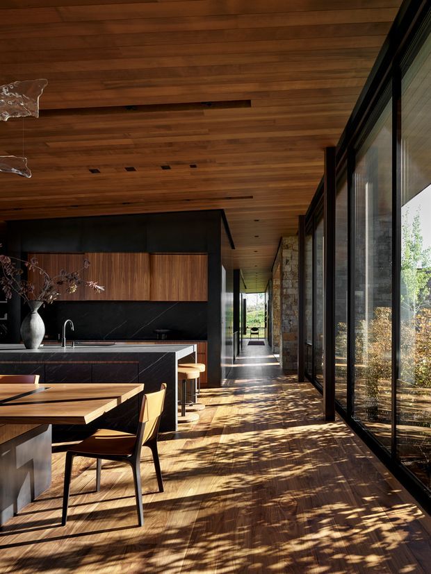 A warm, contemporary open-plan kitchen and dining area extending along a glazed corridor. The space features a rich timber-lined ceiling and timber flooring, dark stone kitchen surfaces with walnut-toned cabinetry, a dining table with angular wooden chairs, black-framed floor-to-ceiling glazing, and a textured natural-stone wall. Strong sunlight filters through the glazing, casting organic shadows across the floor.
