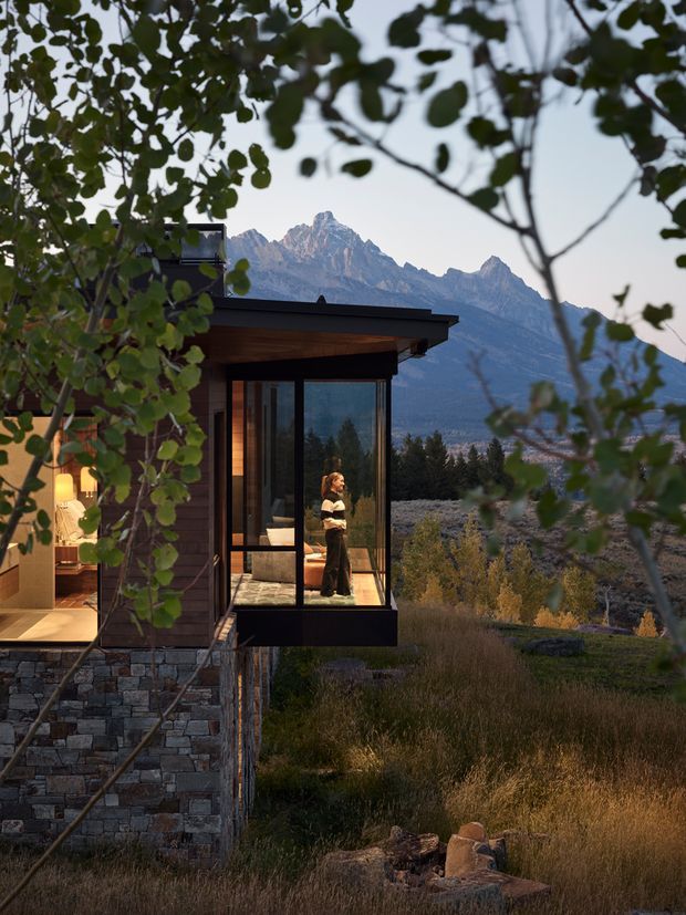 A modern mountain home viewed through leafy branches at dusk. The house features dark timber cladding, a low flat roof, extensive floor-to-ceiling glazing, and a projecting glazed room or bay with a person visible inside. A substantial stacked-stone base anchors the building on the landscaped hillside, with rugged alpine peaks forming the background.