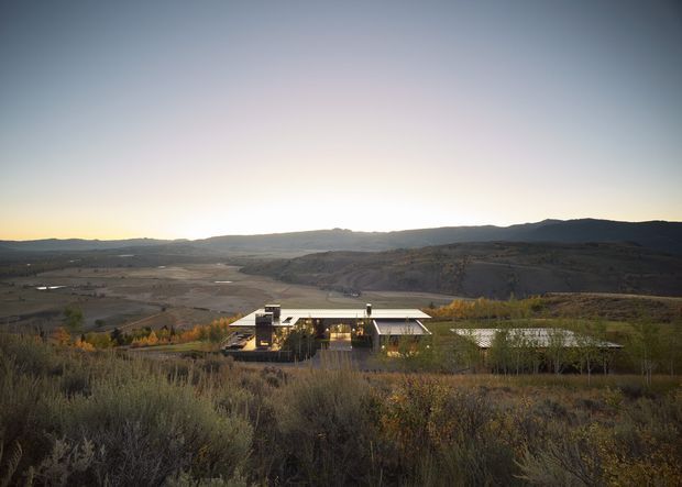 Wide landscape view of a low, contemporary residence set within an expansive arid, shrub-covered rural valley. The house has a predominantly horizontal composition with flat or very low-pitched roof planes, extensive glazing, and warm interior lighting, surrounded by native-looking vegetation and distant rolling hills at sunset.