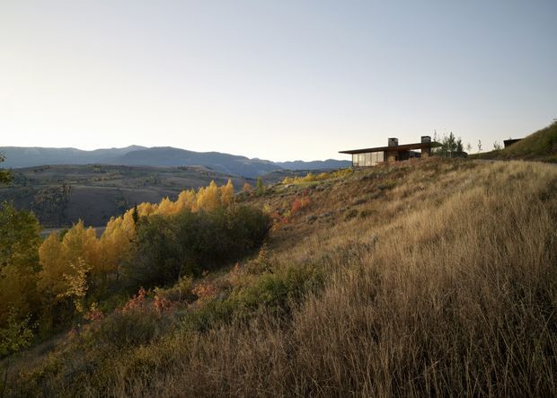A low-profile contemporary house sits on a sloping, grassy hillside overlooking a broad mountainous valley. The building has a long horizontal form, extensive glazing, dark exterior finishes, and a flat or very low-pitched roof. Autumn-coloured trees and dry grasses surround the dwelling in a natural rural landscape.