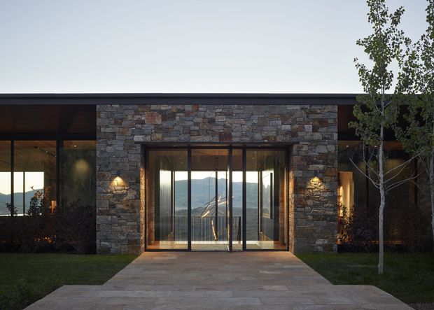 A low, modern stone-clad building facade viewed from a landscaped approach. A broad flat roof and dark-framed floor-to-ceiling glazing surround a central recessed glass entry, with warm interior lighting visible through the windows. The facade uses varied natural stone masonry, and distant mountains are visible through the glazing. A paved path leads across a lawn to the entrance, with slender trees flanking the building.