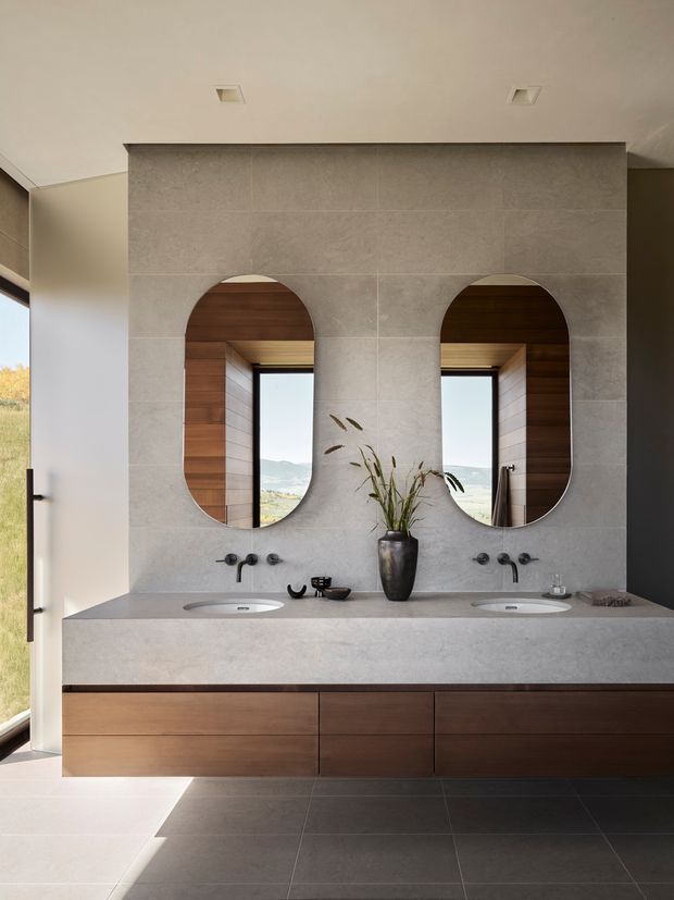 Contemporary double-vanity bathroom with a floating dark timber vanity, grey stone-look tiled wall and benchtop, two integrated basins, black wall-mounted tapware, and paired arched mirrors. Warm timber-lined window reveals and abundant natural light add contrast to the neutral, refined interior.