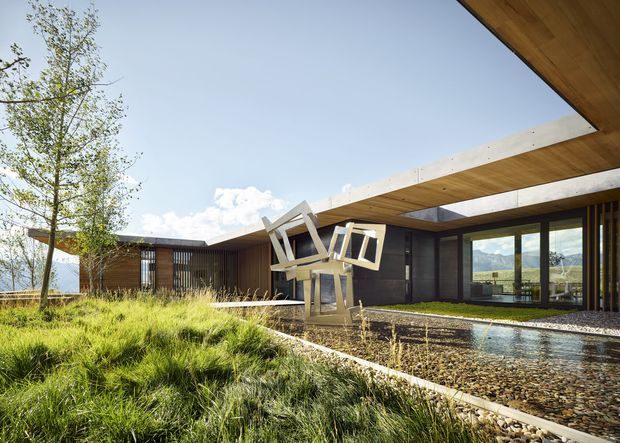 A low-slung contemporary house arranged around a landscaped courtyard and shallow pebble-lined water feature. Broad projecting roof planes with timber soffits, dark glazed walls, vertical timber screening, and exposed concrete roof edges create a strong indoor-outdoor connection. Mountain scenery is visible through the glazing, while sculptural outdoor furniture sits near the water feature and native-style grasses occupy the foreground.