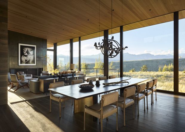 A spacious open-plan dining and living area with a long rectangular dining table, upholstered dining chairs, a sculptural chandelier, and a lounge seating zone beyond. Full-height black-framed glazing provides expansive views of a dry, mountainous landscape. The interior features a warm timber-lined ceiling, dark flooring, dark stone or concrete-toned walls, and restrained contemporary furnishings.
