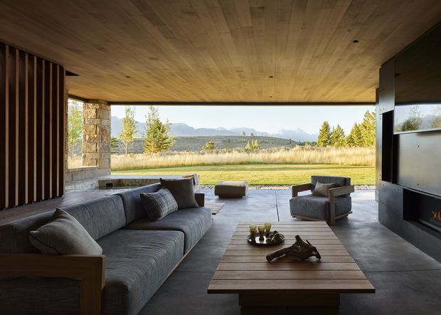 A contemporary open-plan living room with a large sectional sofa, armchair, low timber coffee table, and a built-in fireplace. The room opens broadly to a landscaped rural view and distant mountains through a large glazed opening. Warm timber covers the ceiling, while dark stone or concrete flooring, stone columns, vertical timber screening, and dark metal framing create a refined indoor-outdoor setting.
