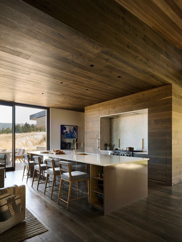 A refined open-plan kitchen with a large island and integrated seating, set within a warm, wood-lined interior. Dark timber flooring, a timber-clad ceiling and walls, pale stone island benchtop, and a recessed stone backsplash create a cohesive natural material palette. Full-height glazing opens to a dry grassland landscape, while minimalist recessed lighting and contemporary cabinetry contribute to the calm, high-end atmosphere.