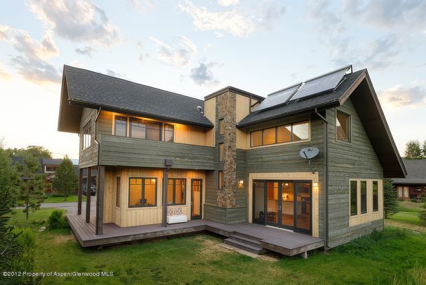 Exterior view of a substantial two-storey timber-clad home with multiple steeply pitched gable roofs, projecting upper-level volumes, dark roof shingles, warm timber-framed glazing, and a central stone-clad vertical section. The house sits within a grassy landscaped residential setting and includes raised timber decks and exterior stairs. Warm interior lighting contrasts with the muted green and natural timber exterior.