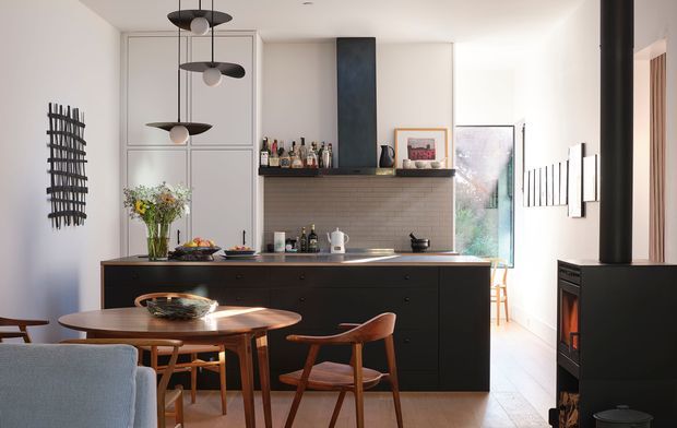 Bright, contemporary open-plan kitchen and dining area with a black island and integrated cabinetry, a tiled backsplash, open shelving with bottles, pendant lights, timber dining furniture, and a dark freestanding wood-burning stove. White walls, timber flooring, and glazing create a warm, refined interior.