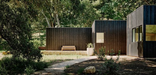 Exterior view of a low, angular contemporary building set within a densely planted wooded landscape. Dark vertical cladding forms several connected volumes, with a large glazed opening at right and a simple bench positioned against the facade. A gravel or concrete path leads through the landscaped foreground.
