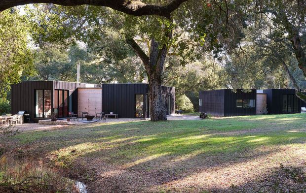 A low, single-storey contemporary residence composed of several dark-clad rectangular volumes set among mature trees. Large glazed openings and warm timber-toned wall sections face a broad grassy landscape, with a small outdoor seating area beside the building. The image is an exterior architectural view in a wooded, secluded setting.