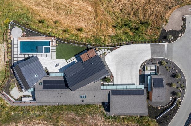 Aerial top-down view of a large contemporary residential property on a sloping site. Several dark-roofed building volumes surround paved terraces and a rectangular swimming pool, with a curved driveway and landscaped planting areas at the right. The site is bordered by rough grassland and retaining edges.