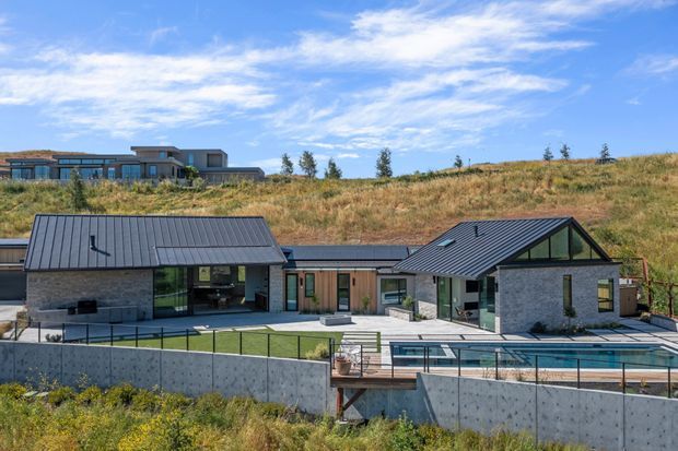 A contemporary single-storey residential property viewed from outside, set into a gently sloping grassy site. The home is composed of several connected wings with dark standing-seam pitched roofs, grey masonry walls, extensive glazing, and timber-clad sections. A swimming pool, paved terrace, concrete retaining walls, and black metal fencing occupy the foreground, while another modern home is visible higher on the hillside.