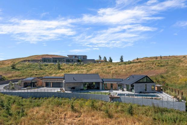 Wide exterior view of a contemporary single-storey residence set into a dry, gently sloping landscape. The home has dark pitched roof forms, light and dark facade sections, extensive glazing, a covered entry or carport area, and a paved outdoor terrace with a pool or water feature at the front. A second modern dwelling is visible on the hillside behind.