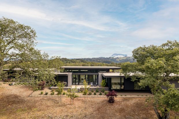 A low-slung single-storey contemporary house set within a dry, landscaped site, viewed from the front. The dark horizontal facade has extensive glazing, a deep shallow roofline, and a central glazed entry or living area. Mature trees frame the property, with a wooded ridge and distant mountain visible beyond.