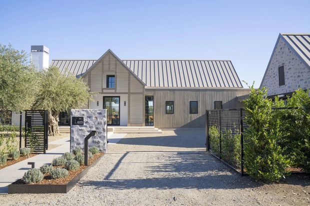Exterior view of a contemporary single-storey residence with a long gabled form, steep metal standing-seam roof, vertical timber-style cladding, dark-framed windows, and a smaller stone-clad gabled volume at right. The property is approached via a gravel driveway and landscaped entry path with low planting, fencing, and mature trees.