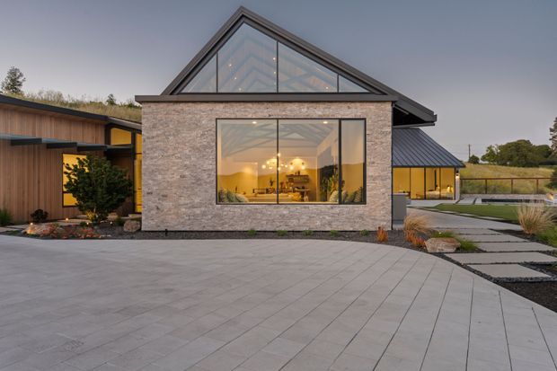 Exterior dusk view of a contemporary single-storey residence with a prominent gabled glazed facade, textured masonry walls, dark metal roof elements, and warm interior lighting visible through large windows. A broad paved driveway and landscaped paths surround the house.