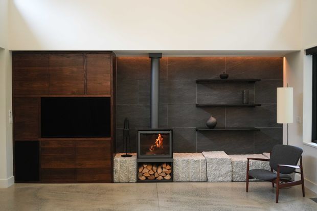 A refined living room with a built-in dark timber media wall and cabinetry, a central wood-burning fireplace with log storage, and a dark tiled feature wall with floating display shelves. A sculptural armchair, floor lamp, firewood, and textured hearth seating create a warm, contemporary atmosphere. The room has a high, softly lit ceiling and polished concrete flooring.