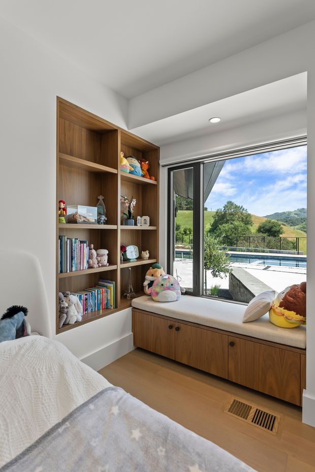 Bright, calm children's bedroom with a built-in walnut-toned shelving unit filled with books and toys, a matching window seat with integrated drawers, and a large glazed opening overlooking a landscaped pool area and rolling green hills. The room has white walls and ceiling, light timber flooring, soft neutral upholstery, and playful children's furnishings.