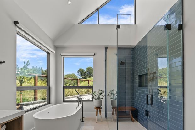 Bright contemporary bathroom with a freestanding white bathtub, glass-enclosed walk-in shower, grey-blue tiled feature walls, white painted walls, and large windows overlooking greenery. A high angled ceiling with clerestory glazing brings in abundant natural light, while timber-framed outdoor views and potted plants add warmth.