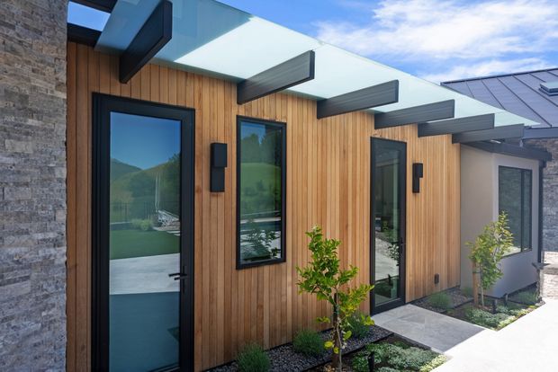 Contemporary single-storey residential facade with warm vertical timber cladding, dark-framed glazed doors and windows, and a series of projecting flat roof or canopy elements. A contrasting stacked-stone wall frames the left side, while a pale rendered section and metal roof are visible to the right. The entry walkway and restrained landscaping complete the modern exterior composition.