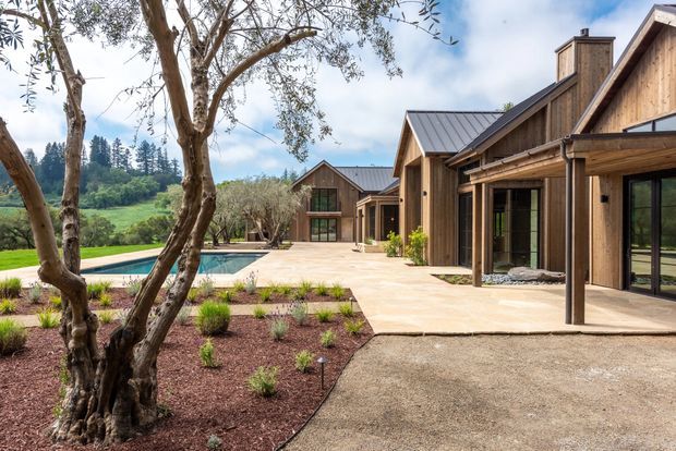 A contemporary timber-clad residential retreat arranged around a broad paved courtyard and swimming pool. The buildings feature repeated pitched gable roofs with dark metal roofing, large glazed openings, covered verandas, and warm natural wood façades. A landscaped garden with mature trees and low planting frames the foreground, while rolling green countryside is visible beyond.