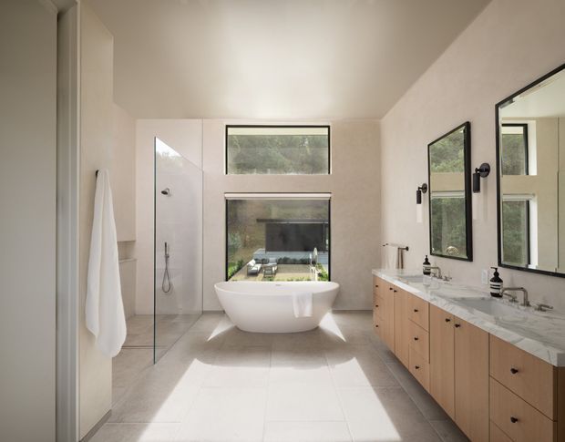 Bright, spacious contemporary bathroom with a freestanding white bathtub positioned in front of large windows, a frameless glass shower enclosure to the left, and a long double vanity with pale timber cabinetry and a light stone benchtop to the right. Black-framed mirrors and fixtures provide contrast, while abundant natural light creates a calm, refined atmosphere.