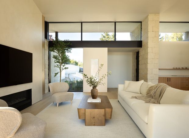 Bright, refined contemporary living room with a large cream sectional sofa, sculptural lounge chairs, a low solid-wood coffee table, and a wall-mounted television above a dark fireplace. Full-height glazing and clerestory windows bring in natural light, while pale textured masonry and plaster walls frame an open connection to a compact timber-fronted kitchen area.