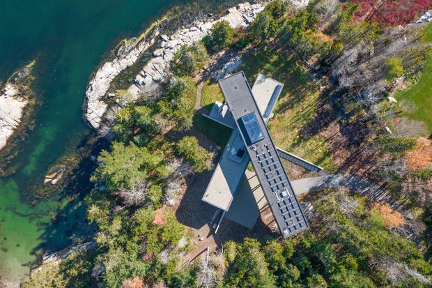 Aerial top-down view of a distinctive angular, multi-wing building nestled among dense woodland on a rocky shoreline beside clear green-blue water. The structure has several pitched roof forms and a long glazed or skylit central connecting section, with paths and natural vegetation surrounding it.