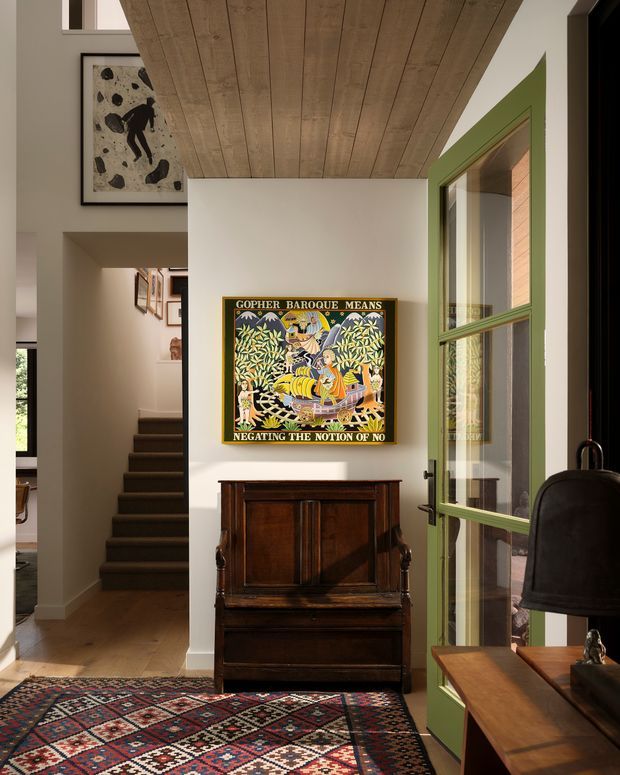 A refined residential entry hall with a dark timber staircase, white walls, a richly patterned geometric rug, framed artwork, and an antique dark-wood console or cabinet. A green-painted glazed internal door opens toward another room, while a timber-lined ceiling creates a strong architectural feature overhead. Warm daylight and layered vintage furnishings give the space an eclectic, inviting character.