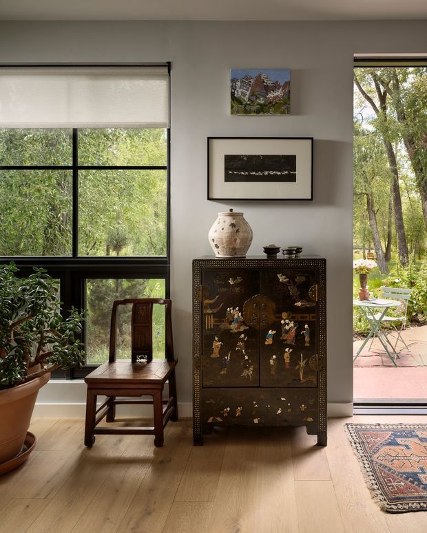 A refined interior vignette with a dark decorative Asian-style cabinet, carved wooden chair, ceramic vessel, framed artwork, and potted plant. Pale timber flooring and warm neutral walls surround large black-framed windows and a glazed door opening to a leafy garden and paved terrace. The room has abundant natural light and a collected, elegant character.