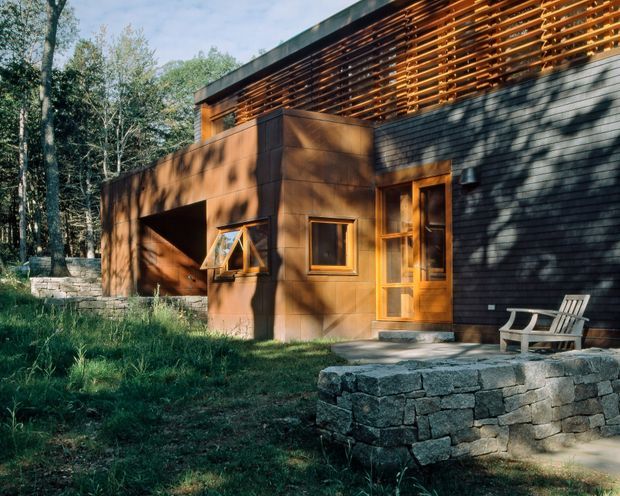 Exterior view of a modern woodland house with a warm timber-clad lower volume, dark shingle-clad upper wall, and a prominent horizontal timber louvre screen beneath a flat roof edge. Timber-framed windows and a glazed entry door face a landscaped garden with stacked-stone retaining walls and a small outdoor bench.