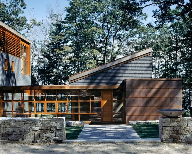 Exterior view of a contemporary, low-rise wooded residence composed of interconnected volumes. The building features dark horizontal shingle-like cladding, warm timber-framed glazing and screens, a sloping roofline, and natural stone retaining walls framing the paved entry path. Mature trees surround the property.