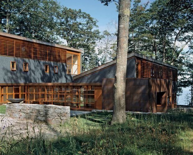 Exterior view of a contemporary multi-volume house set among mature trees. The building features dark horizontal cladding, warm timber window frames and extensive timber screening, with low-pitched roof forms and a stone retaining wall in the foreground.