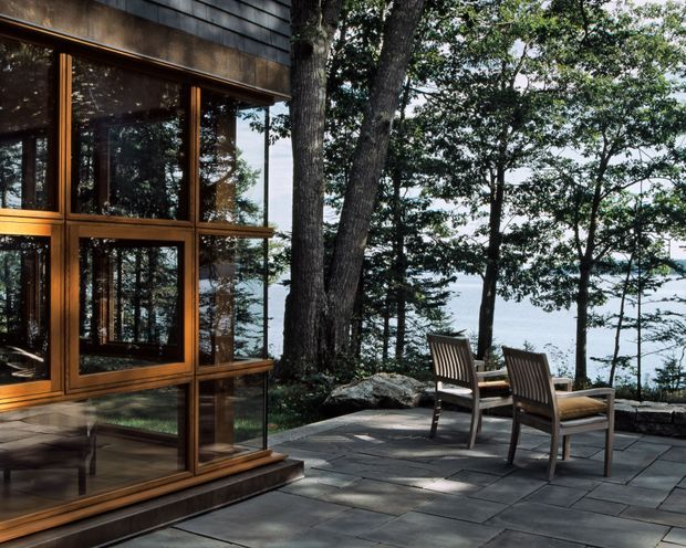 Exterior view of a modern lakeside or waterfront home with extensive floor-to-ceiling glazing framed in warm timber. A paved terrace with outdoor chairs overlooks the water through mature trees, while dark horizontal exterior cladding and exposed rock reinforce the natural setting.