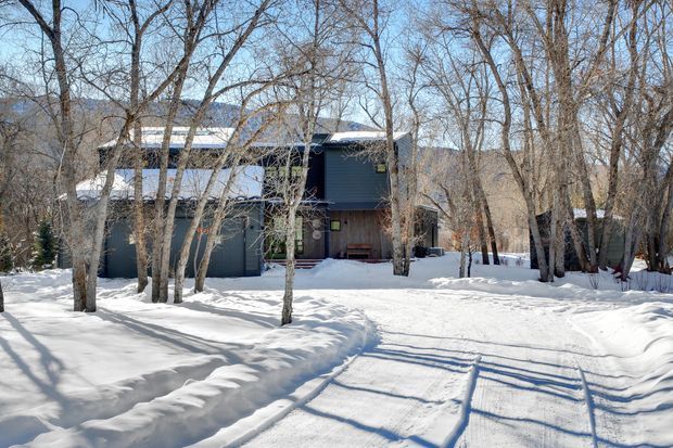 A modern multi-level house set within a snowy, wooded landscape. The dark exterior includes broad glazing, horizontal cladding, and a projecting upper volume, while a cleared driveway and paths lead through the snow-covered site.