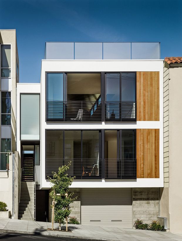 Street-facing view of a narrow multi-storey contemporary townhouse or urban residence. The facade is composed of white rendered walls, large dark-framed glazed openings with horizontal metal balcony rails, vertical timber feature cladding panels, a rooftop glazed screen, and a lower-level integrated garage. Adjacent dense urban buildings and a steeply sloping street reinforce the compact city setting.