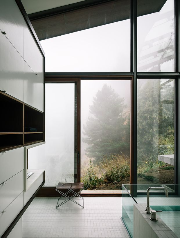 A minimalist contemporary kitchen interior with floor-to-ceiling glazing overlooking a misty, planted landscape. The room features white built-in cabinetry with dark open shelving, a tiled floor, a glass-sided island or peninsula with a tiled base, and a streamlined stainless steel tap. Dark-framed glazing and timber-framed doors create a strong architectural contrast, while a compact stool sits near the window.