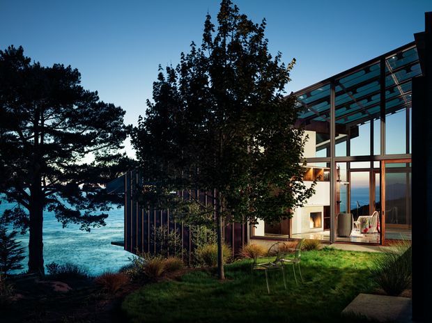 Dusk exterior view of a contemporary waterfront house set among mature trees and landscaped grass. The building features extensive floor-to-ceiling glazing, slim dark framing, warm timber-toned structural elements, and a transparent roof or covered terrace structure. Interior lighting glows through the windows, with the ocean clearly visible beyond the property.