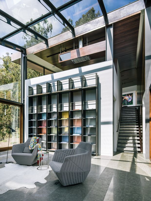 A tall, light-filled contemporary interior with a dramatic glazed roof and floor-to-ceiling windows overlooking trees. The double-height room features dark metal-framed shelving, a timber-lined ceiling and suspended kitchen or display joinery, a polished grey floor, sculptural grey lounge chairs, and an open stair leading to an upper level. Strong daylight creates a refined, gallery-like atmosphere.