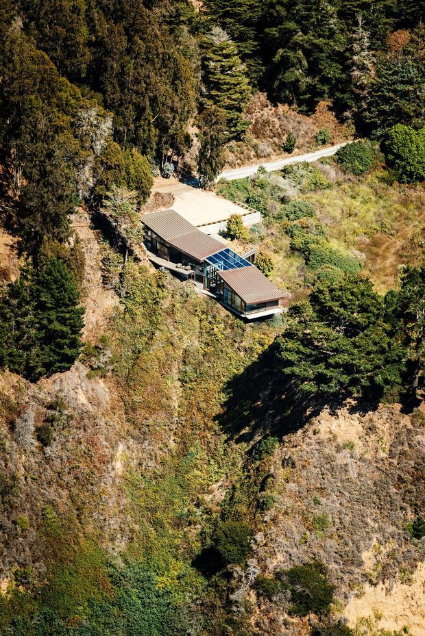 Aerial view of a low-profile modern house embedded in a steep, densely vegetated hillside. The elongated building has dark metal pitched roof planes, extensive floor-to-ceiling glazing, and a central glazed link or courtyard-like section. A driveway and narrow road are visible above the house, while the surrounding rugged terrain is covered with mature trees and scrub.
