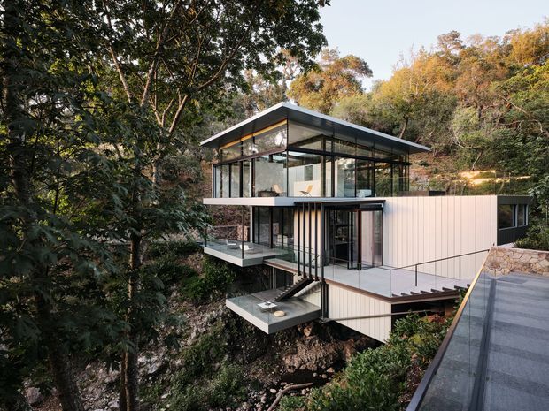 Exterior view of a contemporary multi-level hillside house set among dense trees. The home features extensive floor-to-ceiling glazing, projecting cantilevered balconies, slim dark metal framing, pale vertical wall cladding, and a broad low-pitched roof. A stair and walkway connect the elevated levels to the surrounding landscape.