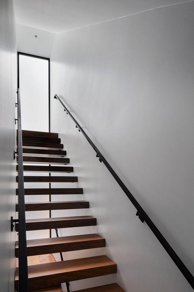 A minimalist modern stairway viewed from below, featuring floating dark timber treads, slim black metal stringer and handrails, and crisp white walls. A tall narrow glazed opening or door brings light into the stairwell, creating a high-contrast, sculptural composition.