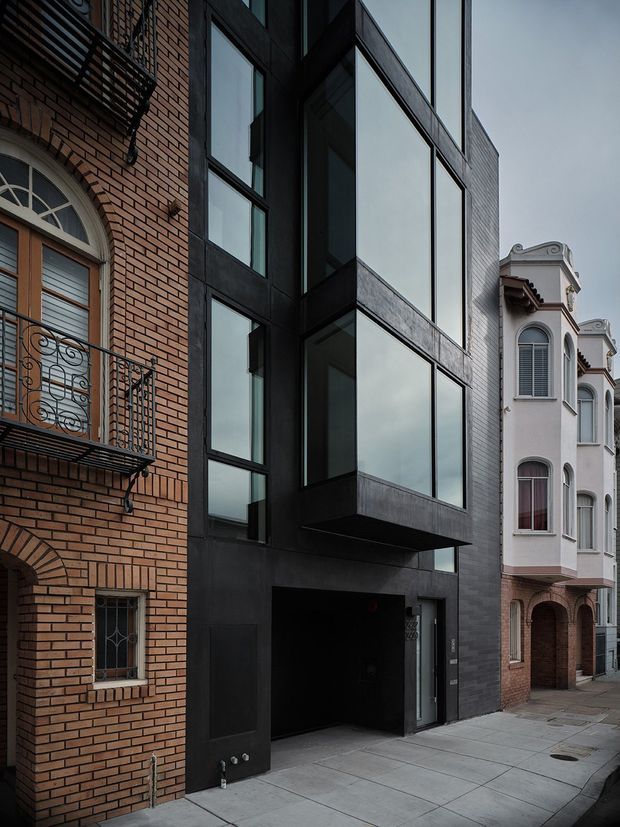 Street-level architectural photograph of a narrow urban streetscape, centred on a contemporary multi-storey building with a dark cladding facade, projecting box-like upper volume, and large black-framed glazed windows. Traditional brick and ornate residential facades directly adjoin it, creating a strong contrast between modern and historic architecture. A recessed ground-level garage or entry opening fronts the concrete-paved sidewalk.