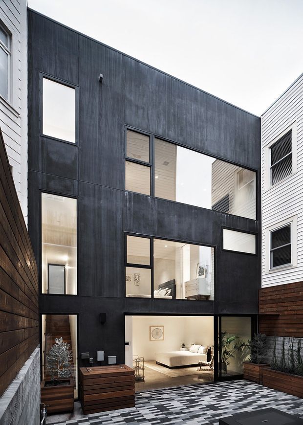 Exterior view of a narrow multi-storey contemporary residence enclosing a private courtyard. The facade is predominantly dark charcoal vertical cladding with large black-framed windows, contrasted by adjacent light horizontal weatherboard walls. The ground-level glazing reveals a bright living room, while the courtyard has patterned paving, timber planter boxes, and outdoor furniture.