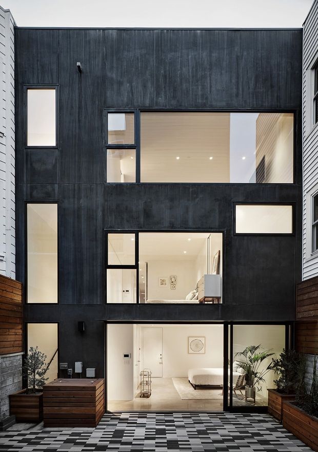 A narrow multi-storey contemporary residence viewed from a small courtyard. The facade is clad in dark vertically textured panels with irregularly arranged black-framed windows and large glazed openings revealing bright minimalist interiors. Timber privacy fencing, raised planter boxes, and a striking geometric black-and-white tiled courtyard occupy the foreground. A bedroom and circulation spaces are visible through the ground-floor glazing.