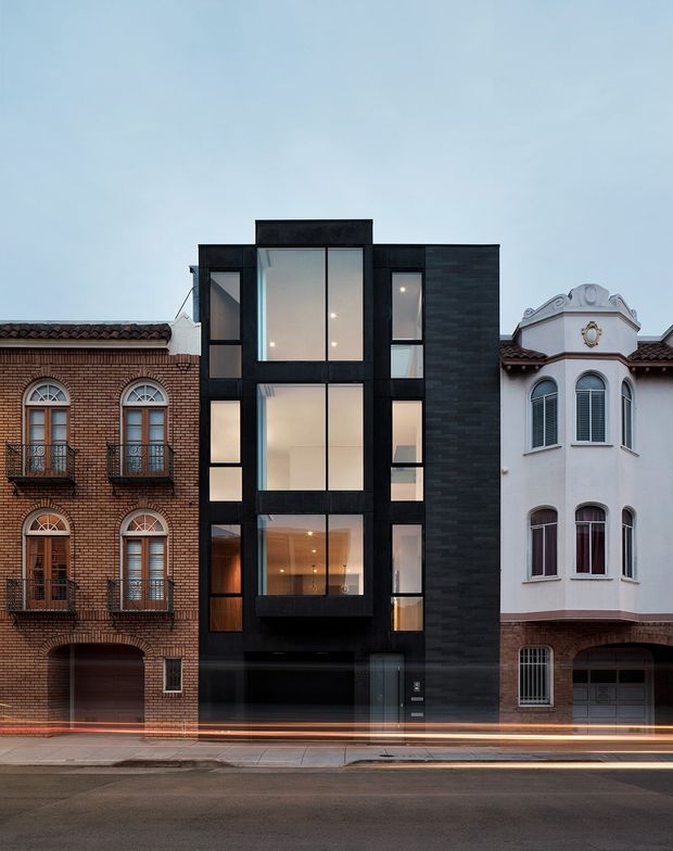 Street-facing view of a narrow contemporary multi-storey infill residence between two older ornate brick and stucco townhouses. The central facade is clad in dark charcoal masonry or paneling and organized around tall stacked floor-to-ceiling windows, with a projecting glazed upper-level bay, recessed entry, and dark garage door. Warm interior lighting and long-exposure light trails appear at street level. The neighboring heritage-style facades provide strong architectural contrast.