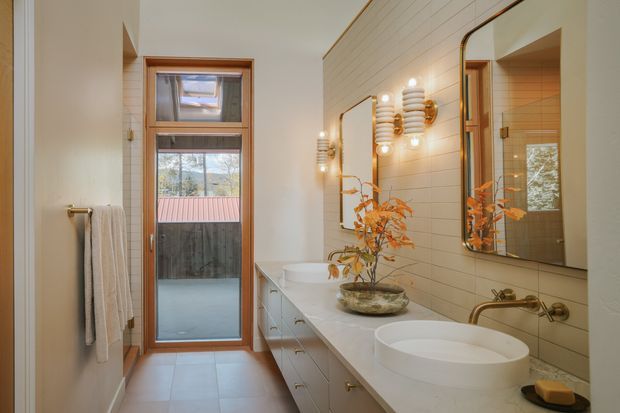 A bright, narrow contemporary bathroom with a long floating vanity, two round vessel basins, wall-mounted brass-toned taps, a large mirror, and twin decorative wall lights. The room features warm timber-framed glazing and door, pale horizontal wall tiles, light stone-look benchtops, and large pale floor tiles. A towel rail and folded towels are visible beside the glazed door.