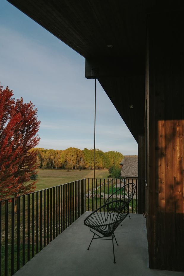 A narrow covered balcony or terrace extends along a dark timber-clad building, with a smooth concrete floor, black metal balustrade, and two wire-frame lounge chairs. The space overlooks an open rural field and autumn trees, with a strong triangular roof overhang creating a sheltered, contemporary outdoor setting.