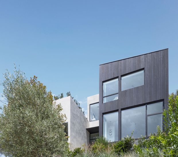 Exterior architectural view of a contemporary multi-level residence, featuring a dark vertical timber-clad upper facade, expansive dark-framed glazing, and contrasting smooth light-rendered walls. The building is partially screened by established landscaping beneath a clear blue sky.