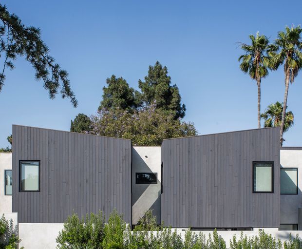 Exterior view of a contemporary single-storey residential building with two projecting, vertically clad grey volumes and a recessed central entry or passage. Dark-framed rectangular windows punctuate the facade, with white rendered walls visible between and around the timber-clad forms. Low landscaping and tall palm trees surround the building under a clear blue sky.